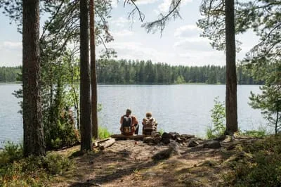Two people sitting on rocks at the edge of a lake, seen through the pines