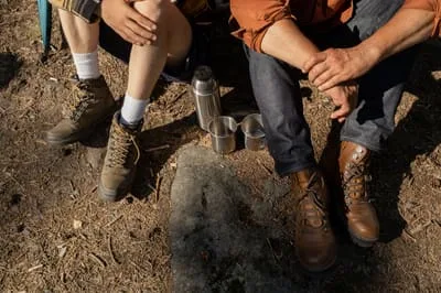 Two pairs of hiking boots beside a thermos on the forest floor